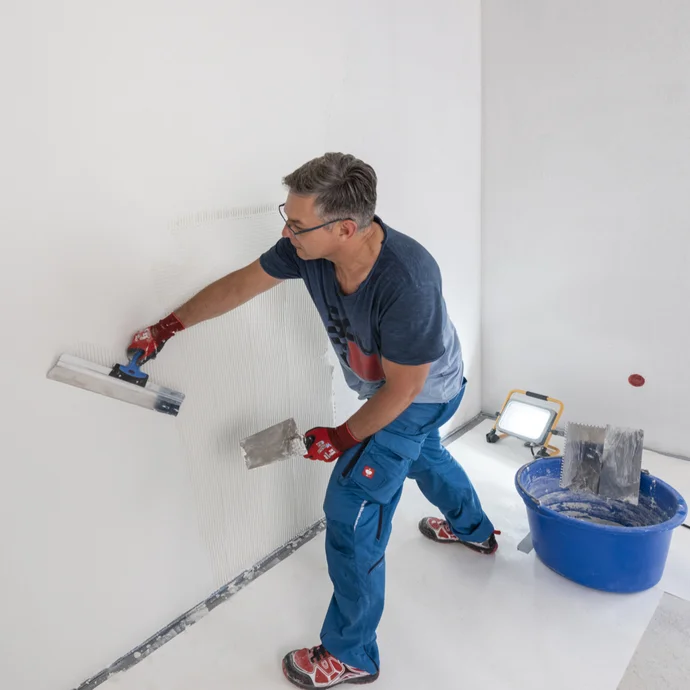 Heimwerker zieht im Raum einen wei&szlig;en, feinen Oberputz eben mit einem Fl&auml;chenspachtel ab. Um Unebenheiten zu sehen, wird die Wand von einem Baustrahler angestrahlt.