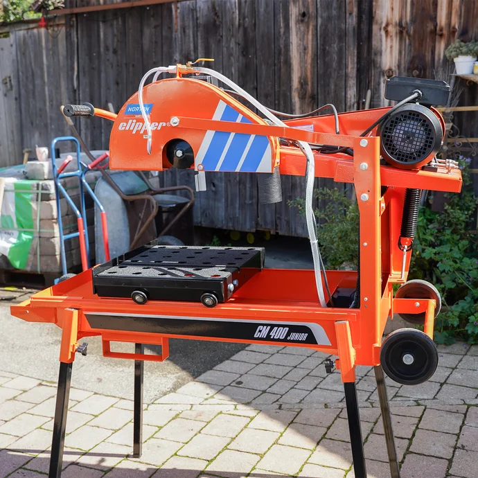 Orangefarbene Steinsäge von Norton Clipper mit Wasserzufuhr  im gepflasterten Hof zum präzisen Schneiden von Beton- und Natursteinen – ideal für Heimwerkerprojekte im Garten- und Landschaftsbau.
