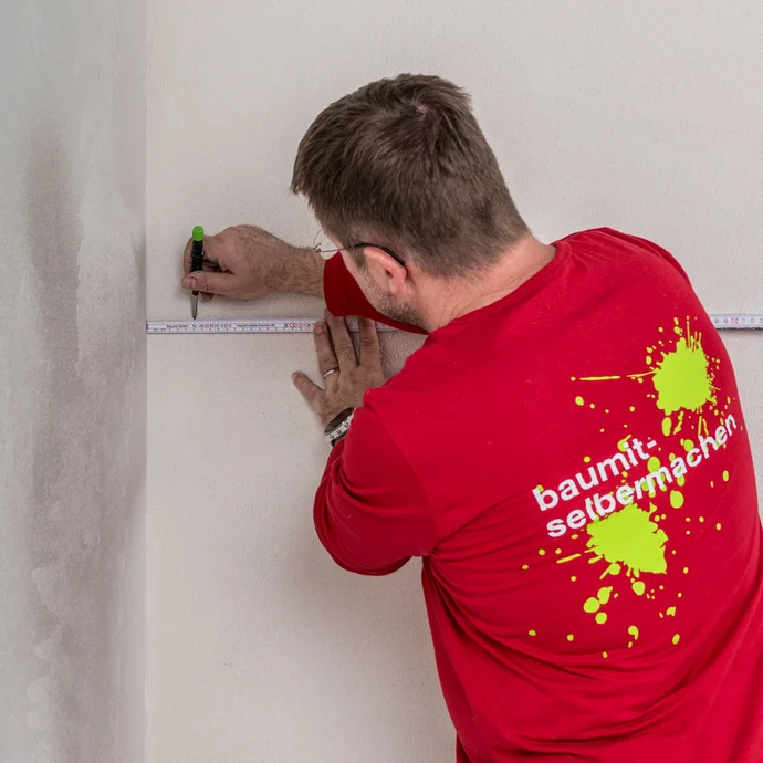 Heimwerker markiert mit Zollstock und Stift die richtige Position f&uuml;r das Baumit Fugendichtband im Sto&szlig;bereich an der Innenwand &ndash; Markierungspunkte setzen f&uuml;r eine b&uuml;ndige Verklebung der Holzfaserd&auml;mmplatten, Baumit Selbermachen Anleitung.