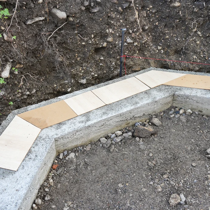 Selbst angefertigte Schablonen für die Mauersteine aus MDF-Platten liegen auf dem Fundament, um die Mauersteine richtig zuzusägen. Eine rote Richtschnur, die am Erdnagel befestigt ist, ist über dem Fundament gespannt.