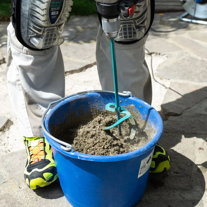 Person rührt mit einem elektrischen Rührwerk Steinkleber Trass in einem blauen Eimer an.