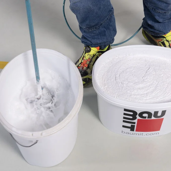 Rührquirl wird in einem Eimer Wasser vorgereinigt. Daneben steht ein pastöser Strukturputz im Baumit-Eimer.