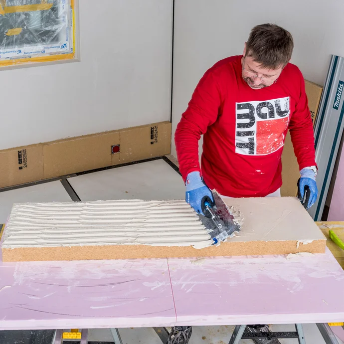 Heimwerker tr&auml;gt Baumit Klebem&ouml;rtel vollfl&auml;chig auf Holzfaserd&auml;mmplatte auf und zahnt die Fl&auml;che mit einer Zahnkelle auf &ndash; vollfl&auml;chige Verklebung der Innend&auml;mmung ohne Hohlr&auml;ume sicherstellen, Baumit Selbermachen Anleitung.