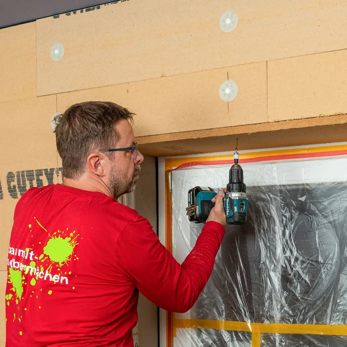 Heimwerker verschraubt die angeklebte Laibungsplatte aus Holzfaser am Sturz des Fensters. Innendämmung mit Tragkonstruktion selbst bauen. Baumit Anleitung zum Selbermachen Schritt für Schritt erklärt.