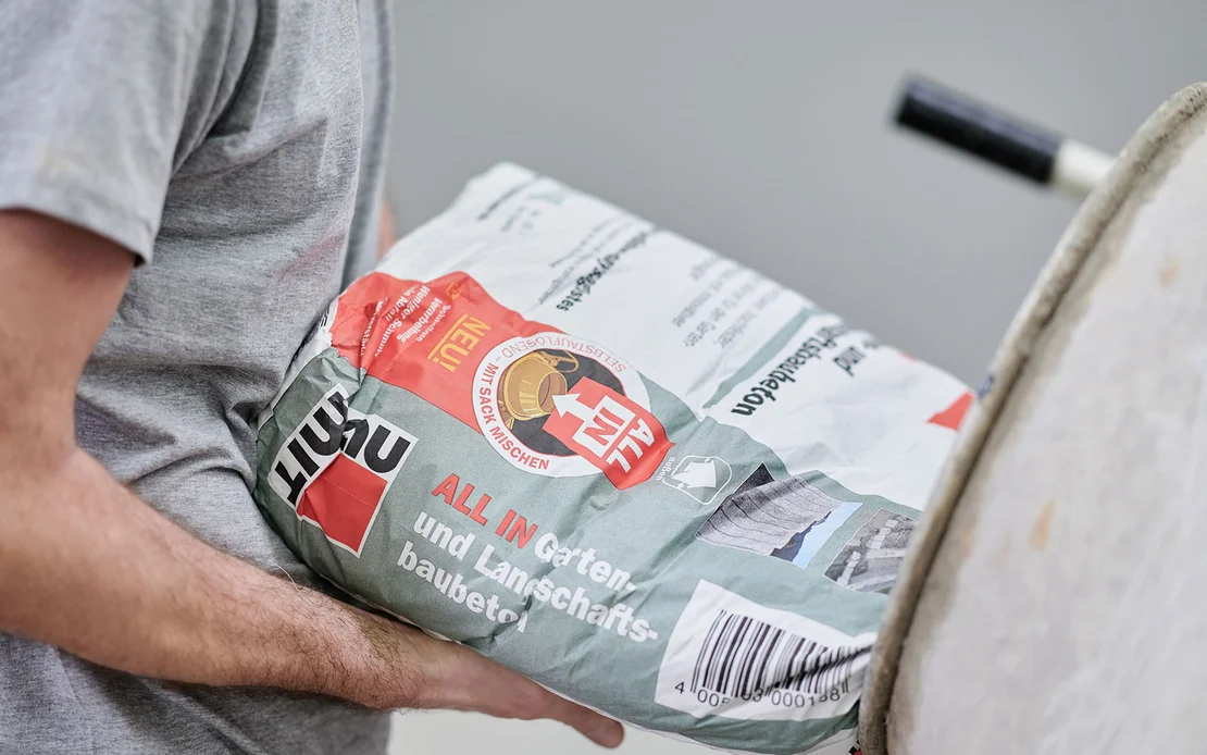 Person mischt Baumit Zementm&ouml;rtel f&uuml;r Heimwerkerprojekte, h&auml;lt Sack neben Betonmischer bereit. Ideal f&uuml;r Garten- und Landschaftsbau.