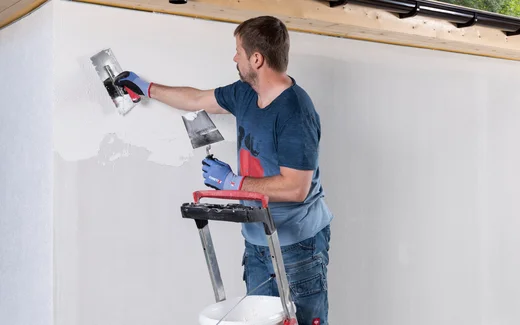 Person steht auf Leiter und tr&auml;gt Strukturputz mit einer Gl&auml;ttkelle an einer Wand auf.