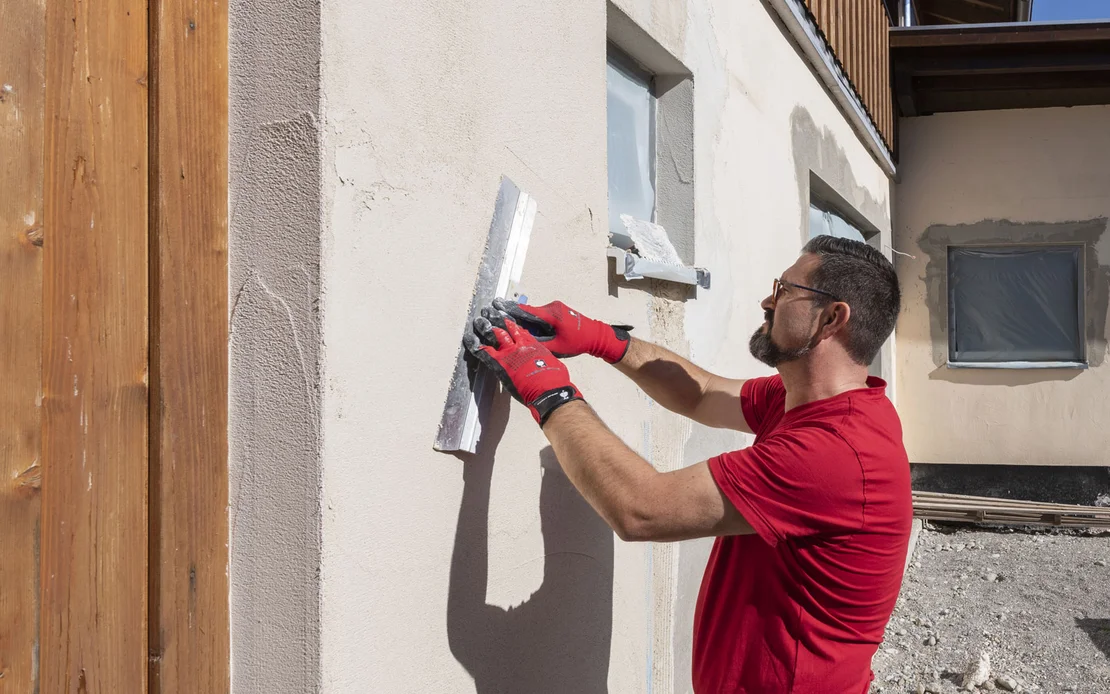 Mann gl&auml;ttet den aufgetragenen Putz mit einer Fl&auml;chenspachtel an der Fassade.