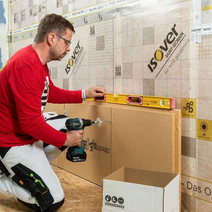 Heimwerker richtet Holzfaserdämmplatte waagerecht aus und verschraubt sie mit Schraubdübeln auf der Holztragkonstruktion. Die Wasserwaage ist zur Kontrolle im Einsatz.