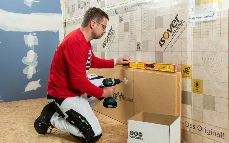 Heimwerker verschraubt die erste Reihe Holzfaserdämmplatten auf der Holztragkonstruktion und kontrolliert die waagerechte Ausrichtung mit der Wasserwaage. Innendämmung selber machen.